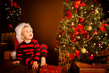 Happy Kid near Christmas Tree, Happy Baby Boy in Night Xmas Room, Child in Red Santa Hat with Present gift box