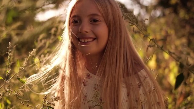 Cute blonde girl plays with her hair and smiles in slow motion backlit with the sunlight. Teenage girl wearing floral dress in the nature during warm summer day