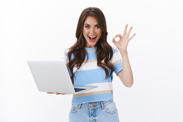 Fototapeta Astonished pretty caucasian woman hold laptop, look camera satisfied give approval, show okay ok sign pleased, rate good application, leave positive feedback good book review, white background obraz