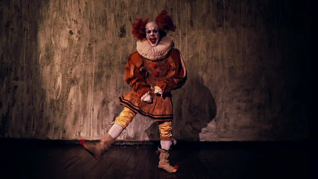 Crazy scary clown dancing against a dark red wall in darkened room. Terrible clown with colorful makeup in a festive costume. Shooting in slow motion