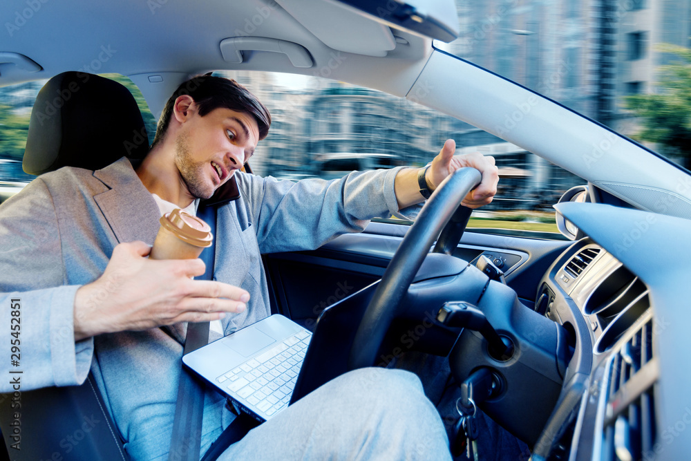 Wall mural man is working on laptop, talking on phone, drinking coffee and driving car. businessman in grey sui