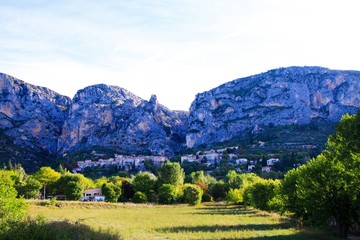 Fototapeta na wymiar View over green valley on French village with giant mountain face background in early morning - Moustiers Sante Marie, Provence, France