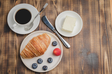 Coffee cup, croissant with berries in white bowl and butter knife on wooden table. Top view and copyspace. Healthy breakfast with fresh berries