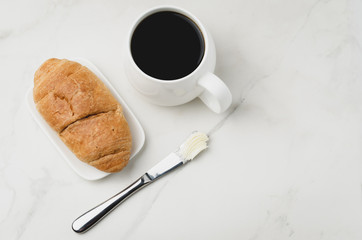 French breakfast. White mug with black coffee and croissant with butter knife on white stone table. Top view, copyspace. Coffee break.
