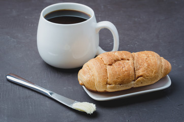 French breakfast. White mug with black coffee and croissant with butter knife on dark stone table. Coffee break.