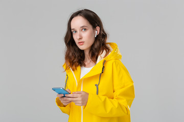 Brunette woman in a yellow raincoat is reading notification on the smartphone. Model wears wireless earbuds. Concept of communication.