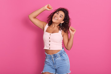 Horizontal shot of gorgeous brunette lady wearing rosy top and jeans, posing isolated over pink background. Charming girl dancing with hair waving, keeps hands up, looking directly at camera.
