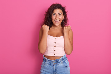 Photo of happy young woman with beautiful dark wawy hair screaming and clenching fist while looking directlyat camera, wearing stylish outfit, isolated over pink background. People emotions concept.