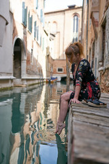 Caucasian redhead woman with floral dress sitting near a canal in Venice barefoot
