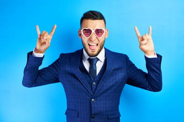 Cool asian kazakh businessman in blue suit raising up hands with rock-n-roll gesture isolated in...