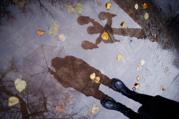 Girl in a coat, black shoes and with an umbrella. Autumn weather in the city, rain, reflection on the pavement. Drops of rain, fallen leaves.