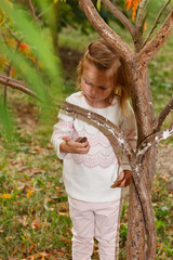 Adorable baby girl holds snail, playing in a sunny park under a tree with yellow leaves, hiding behind tree