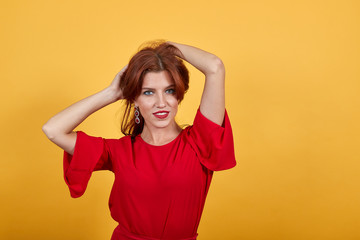 Happy European woman smiling, keeping her hands behind your head. Gorgeous woman in red clothes with reddish lipstick standing over yellow background.