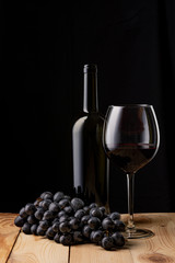 Wine bottle and a glass of wine. A bunch of ripe black grapes. Wooden table. Black background.