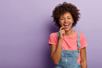 Happy fashionable woman smiles carefree, wears t shirt and denim overalls, keeps fore finger on lips, laughs joyfully during funny conversation, has healthy dark skin, isolated over purple background