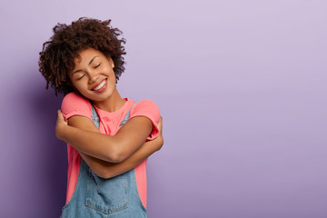 Lovely curly woman embraces herself with pleasure, feels comfort, calmness and love, tilts head and...