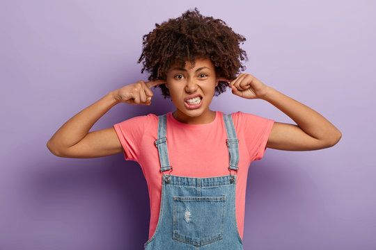 I cannot hear it anymore. Dissatisfied annoyed curly woman keeps index fingers in earholes, ignores annoying sound, clenches teeth, dressed in casual outfit, plugs ears, doesnt listen bad words