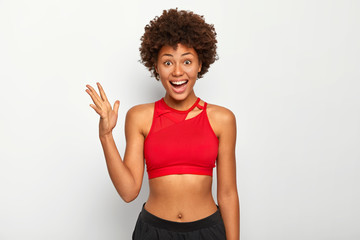 Photo of cheerful dark skinned woman raises palm, feels glad after workout, wears red sport bra,...