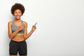Portrait of good looking Afro woman points away on blank space, smiles pleasantly, wears top and...