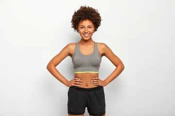 Photo of optimistic dark skinned sporty woman keeps hands on waist, smiles happily, dressed in...