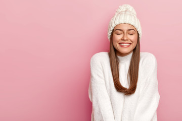 Horizontal shot of pretty young female with dark hair, keeps eyes shut, imagines pleasant moment, smiles pleasantly, shows white perfect teeth, enjoys comfort in new bought sweater, warm hat