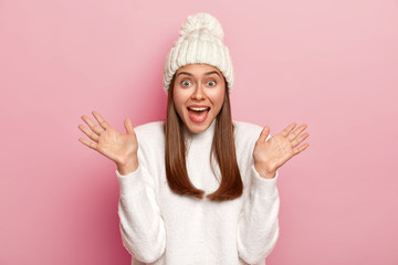 Emotional young Caucasian woman raises palms, keeps mouth opened, has fun, dressed in winter white clothes, gets pleasant surprise, poses over pink background. People, emotions, reaction concept