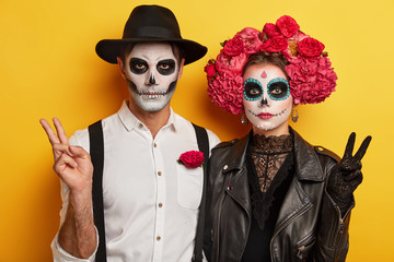 Indoor shot of serious woman and man in special scary costumes, make peace victory gesture, have...