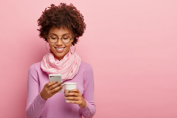 Horizontal shot of cheerful dark skinned girl sends text messages, surfs internet, enjoys free time...