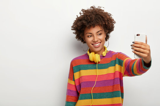 Cheerful dark skinned woman with dark curly hair, makes selfie via smartphone, dressed in colorful sweater, smiles gently, being music meloman, stands against white background, blank space aside