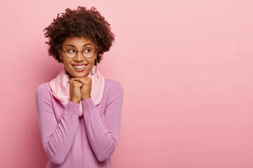 Joyful smiling adorable female keeps both hands under chin, looks aside, dressed in casual clothes, wears spectacles for vision correction, enjoys good news, isolated over pink studio background