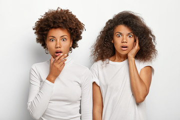 Terrified two sisters with Afro haircut, feel stunned, watch horror movie, wear white casual clothes, being troubled, stand indoor, isolated over white background. People, emotions, facial expressions