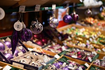 Silver glittered purple Christmas ornaments hanging from shelf in store at Christmas fair in Europe. Blurred background of boxes with colorful glass Christmas baubles