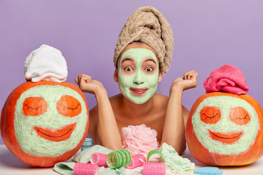 Women, cosmetology and skin care concept. Pleased Asian woman with clay mask, poses topless, raises hands, wears wrapped towel on head, makes funny pumpkins for demonstrating beauty products effect