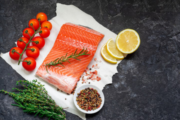 Raw salmon filet with paper with salt, peppers, lemon, thyme and rosemary on over dark stone background, wild atlantic fish . Creative layout made of fish, top view, flat lay