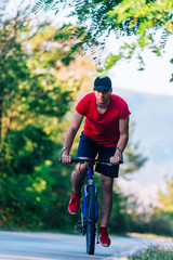 Fit cyclist rides his bicycle (bike) on an empty road in nature wearing a baseball hat and red t-shirt.