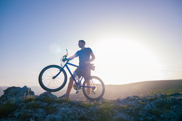Mountain biker holding his bike on a rough cliff terrain on a sunset.