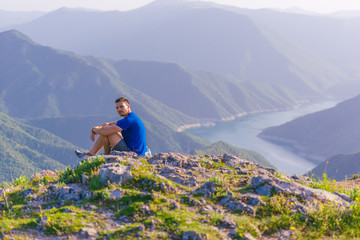 Naklejka premium Adventurous man sitting on top of a mountain and enjoying the beautiful view, while looking downhill at the blue river and amazing mountain line..