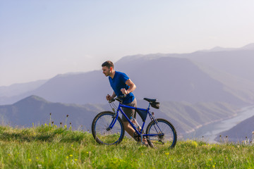 Fototapeta premium Tired fit mountain biker pushing his bike uphill at the top of the mountain on a sunny day with amazing view on a blue river.