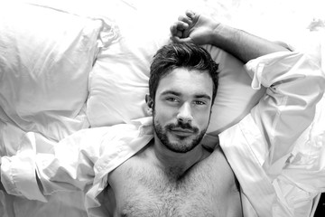 Portrait of handsome hairy muscular man with beard and open white shirt, looking up from bed at...