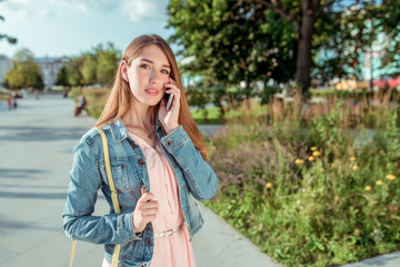 Girl student in pink dress jeans, summer city park, free space, makes phone call, listens to message on mobile phone, online application on Internet. Bag over shoulder.