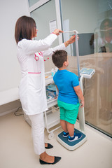 Female doctor measuring the height of little boy in a clinic ( hospital ) while smiling and being...