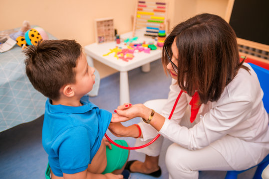 Portrait of adorable little boy visiting doctor, looking brave and smiling, holding while pediatrician listening to heartbeat with stethoscope