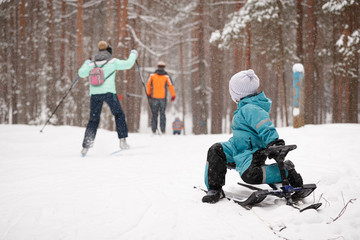A little toddler boy in a blue winter jumpsuit sits on a snow scooter and looks at a family of skiers running through. Child with sleigh in winter park. Lifestyle concept