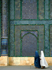 The Blue Mosque in Mazar-i-Sharif, Balkh Province in Afghanistan. Two women wearing burqas (burkas) walk past a wall of the mosque adorned with colorful tiles and mosaics. Northern Afghanistan.