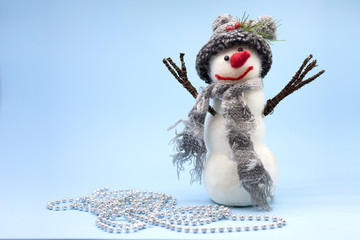 Snowman in gray hat and gray scarf stands near silver beads. Toy snowman on a blue background. Place for text