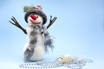 Toy snowman on a blue background. A snowman in a gray hat and a gray scarf stands near a white rat. Year of the white rat. Place for text