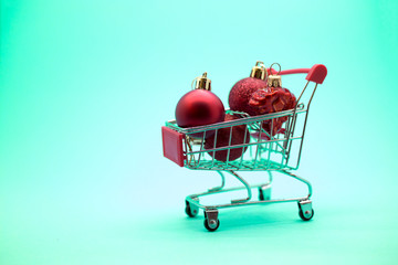 Miniature model supermarket cart with red Сhristmas balls inside on a green backgound. New Year discounts and sales concept
