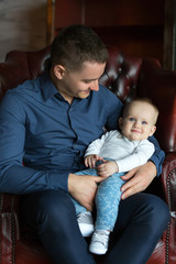 A father sits in a chair and holds his little son in his lap. Man looks at the child. Family life.