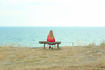 Naklejka na ściany i meble A little girl sits alone. Solo travelling. A tourist sits on a wooden bench on a cliff above the sea coast.