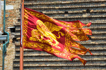 Flag of Venice waving on street in Venice, Italy. Golden winged lion on red flag, closeup view. Old Venetian flag is on background of vintage house wall in summer.